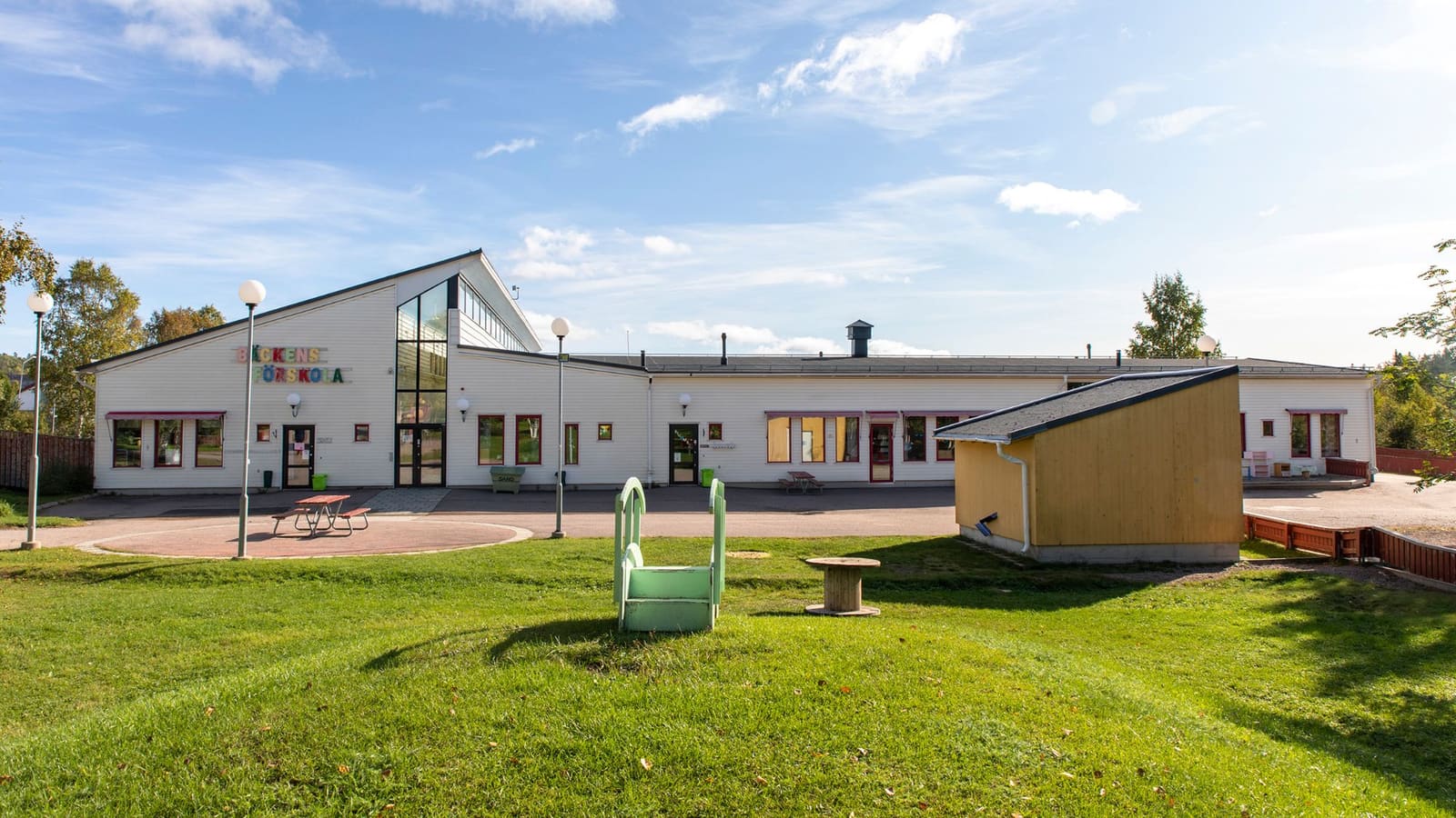 Vit trähusbyggnad med skylten Bäckens förskola en solig sommardag, med grön gräsmatta och blå himmel.